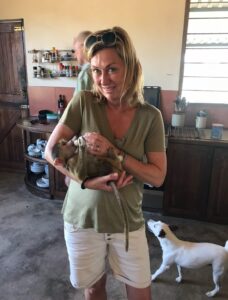 Woman in a green shirt holds a baby monkey inside a kitchen. A dog stands nearby looking up at her.